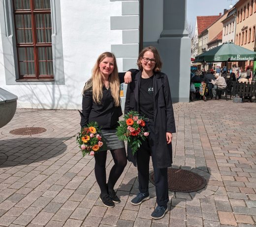Die beiden Sprecher'innen stehen auf dem Marktplatz von Freiberg mit Blumensträußen in der Hand.