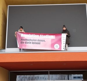 Foto der Banneraktion am Campus der TU Chemnitz. Menschen vom StuRa der TU Chemnitz stehen auf einem Vorsprung über einer Glastür vor einer großen Tafel und halten ein Hochschulgesetzbanner mit "Revolution Studium - Hochschulen denen, die darin lernen!"