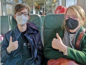 Foto von Maximilian (Beauftragung Koordination) und Fay (Referent*in Feminismus) im Zug mit Maske und Daumen hoch.