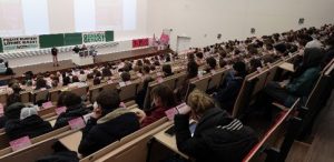Foto der studentischen Vollversammlung in Leipzig, Blick von hinten aus dem Hörsaal auf die Massen an Studierenden. Vorn sind Banner von Genug ist Genug und TVStud zu sehen.